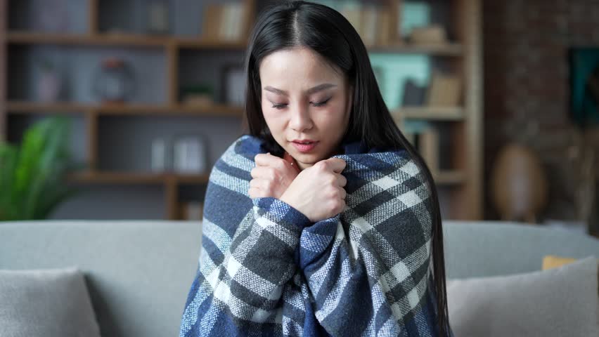Young asian woman is frozen covered with blanket sitting on sofa in living room at home. Female is cold indoors and warms her hands by rubbing. Girl feels discomfort due to low temperature. Close up