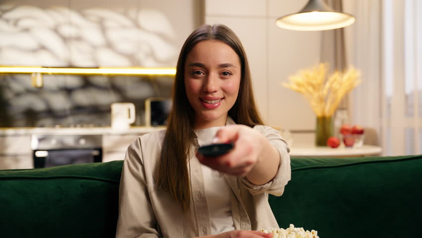 Caucasian woman female lady girl at home relaxing at couch relax at sofa weekend eating popcorn switch channel with remote controller watching TV film movie television program enjoy broadcasting smile