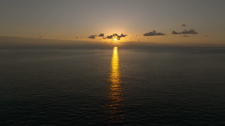 Drone view of a golden pathway on the sea or ocean surface with the sun at sunset or sunrise and a few clouds