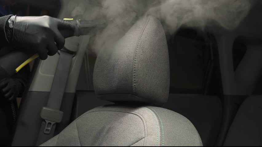 Steam vaping cleaning of car headrest pillow after foam washing with brush 4k video. Worker in car detailing service washing fabric interior of auto using steam generator.