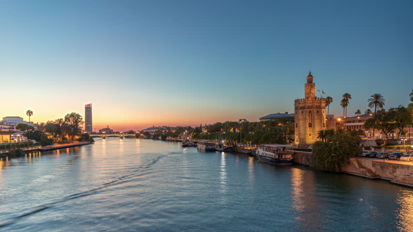 Tour boat floating along the Guadalquivir River near Torre del Oro watchtower in Seville, Spain. Day to night transition timelapse with city lights reflecting on water and scenic view of a waterfront