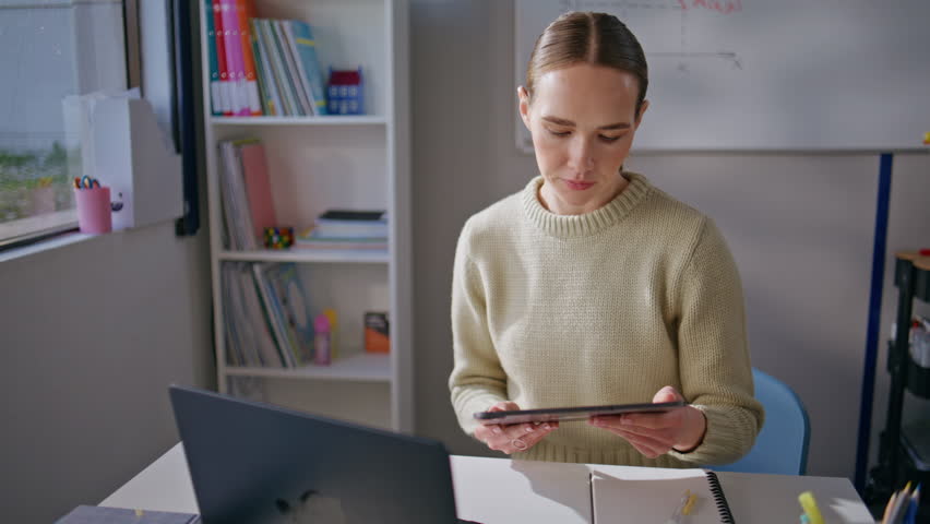 Focused teacher using tablet at school workplace closeup. Calm serious woman working at class room preparing to online lesson. Attentive educator checking homework using pad computer at cabinet alone