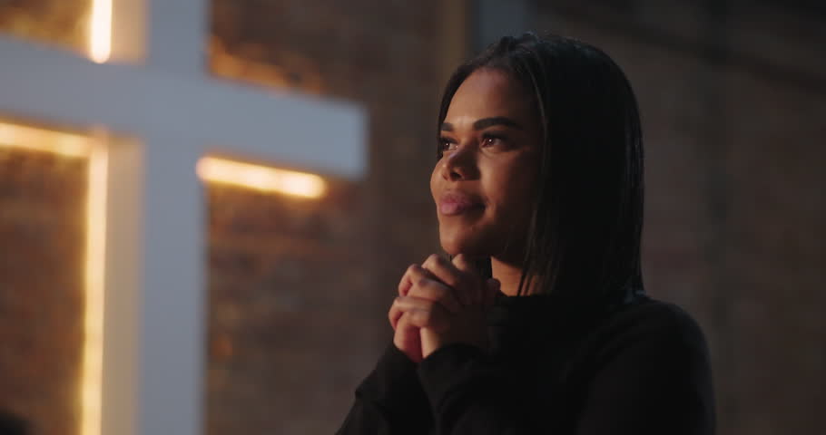 Hispanic woman looking upward in prayer, hands clasped in deep reflection and connection to faith during evangelical church service, glowing cross as background