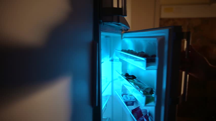 A man opens the refrigerator in the evening and takes food from it