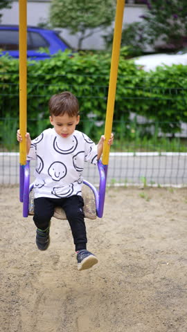 Cute toddler boy sits on the swing. Little kid spends time at playground in summer. Vertical video.