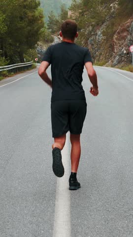 Back follow shot of running muscular athletic man in black sportswear. Sportive male athlete jogging in mountains, preparing for run race competition or marathon. Vertical video