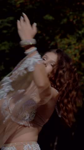 Vertical. Girl belly dancing in the forest. Oriental dance. Woman in white belly dance costume posing in field with flowing movements. Elegant dancer in white costume. White-clad woman showcasing