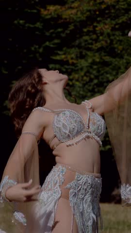 Vertical. Girl belly dancing in the forest. Oriental dance. Woman in white belly dance costume posing in field with flowing movements. Elegant dancer in white costume. White-clad woman showcasing
