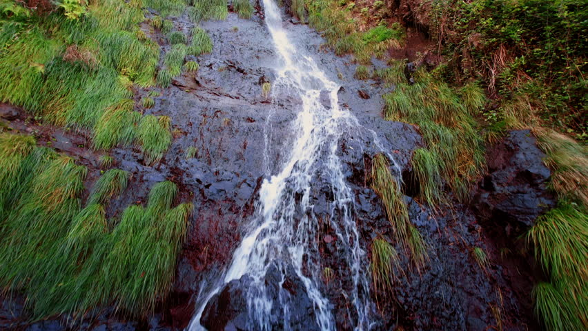 Water flows by the high rock with greenery growing around. Drone footage along the waterscape in Madeira, Portugal.