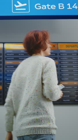 Airport Terminal: Back view of Woman with Luggage and Ticket Checking Flight on Digital Departure Board. Diverse People and Airport Staff Walking In Background. Flight Information Display. Slow Motion
