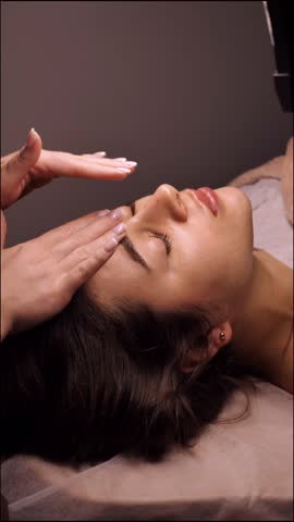 Beautician doing female anti-wrinkle rejuvenating facial massage in beauty salon, close-up, vertical