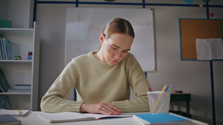 Serious teacher working school closeup. Calm educator in glasses correcting mistakes checking homework at class room. Young woman holding notebook reading attentively at workplace. Occupation concept