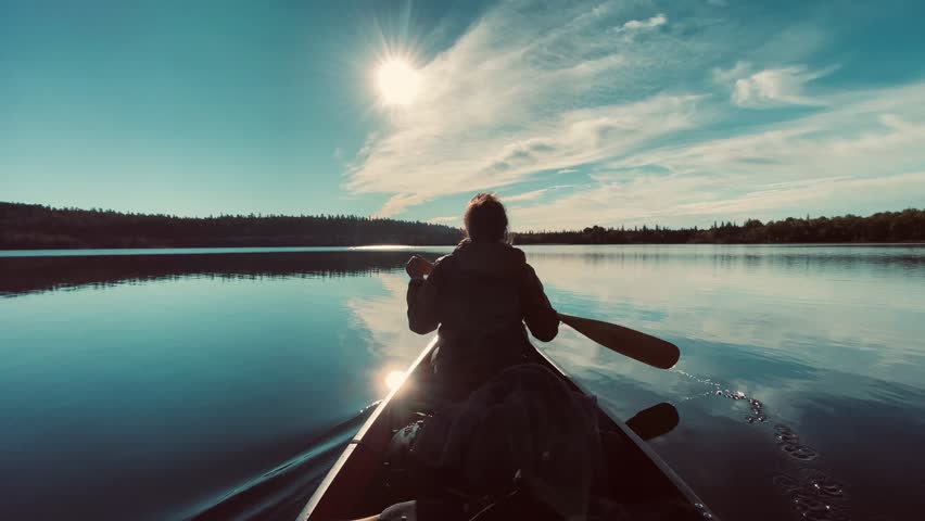 A woman paddles a canoe on a calm lake, surrounded by lush evergreen trees reflecting on the water. The light of late afternoon enhances the tranquil and immersive wilderness experience