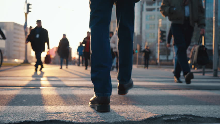 Back view following male feet steps walking pedestrian crossing street on sunny autumn day. Man wearing leather boots goes crossroad. Low angle people legs stepping backlit warm sunlight