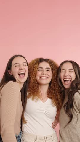 Three young women laughing together on pink background