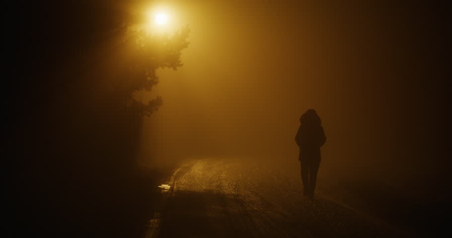The silhouette of a person walking through a dark alley is lit by the distant glow of a city lantern.