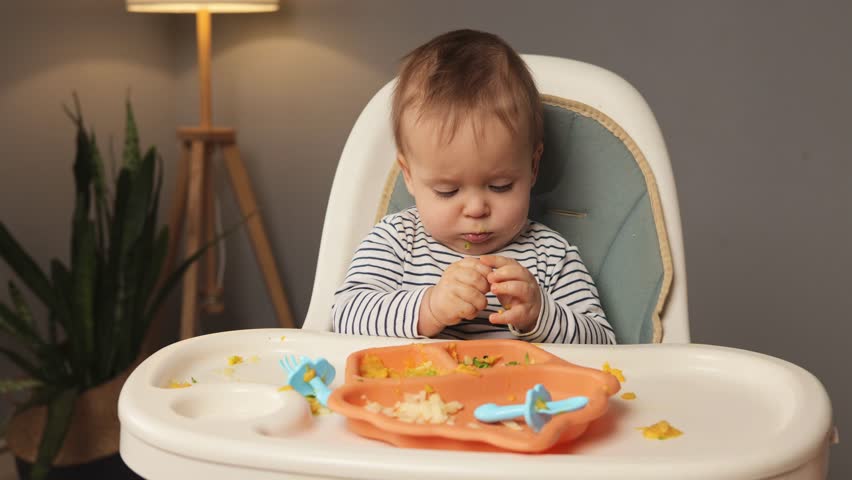 A young child sits in a high chair, curiously playing with food on a bright orange plate. Cheerful messiness surrounds the child, highlighting the joy of self-feeding in a warm setting