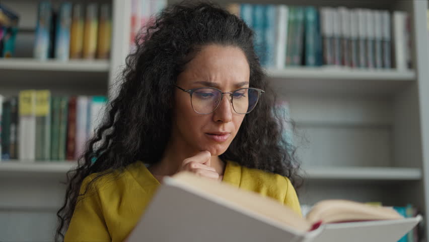 Indian Hispanic woman reader focused preparing exam reading book university library high school girl female student studying read learn textbook literature knowledge glasses smart bookshelf education