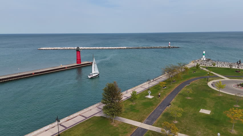 Scenic marina with sailboat and colorful lighthouses on a sunny day in Kenosha, Wisconsin, Sailboat passing by a red lighthouse near a marina surrounded by green park spaces and paved paths.