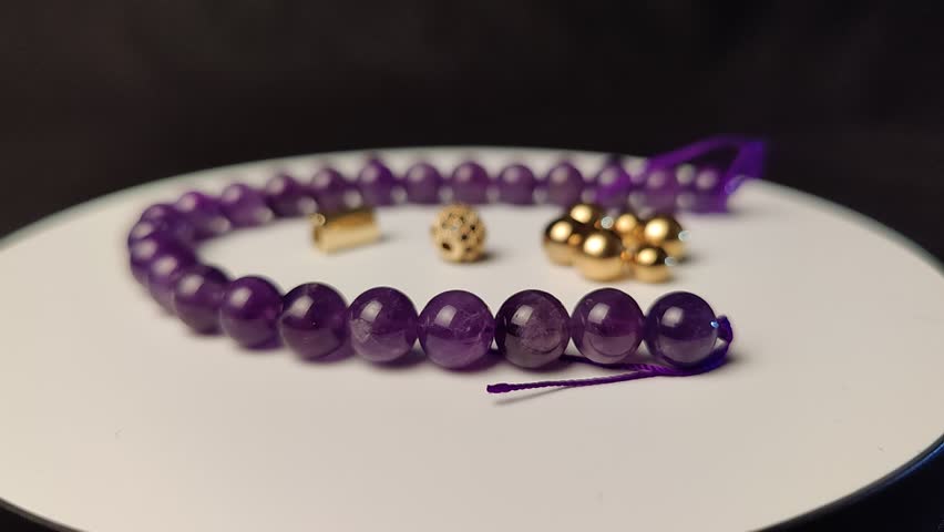 A closeup of a purple Amethyst bracelet and golden jewelry spinning on the white surface