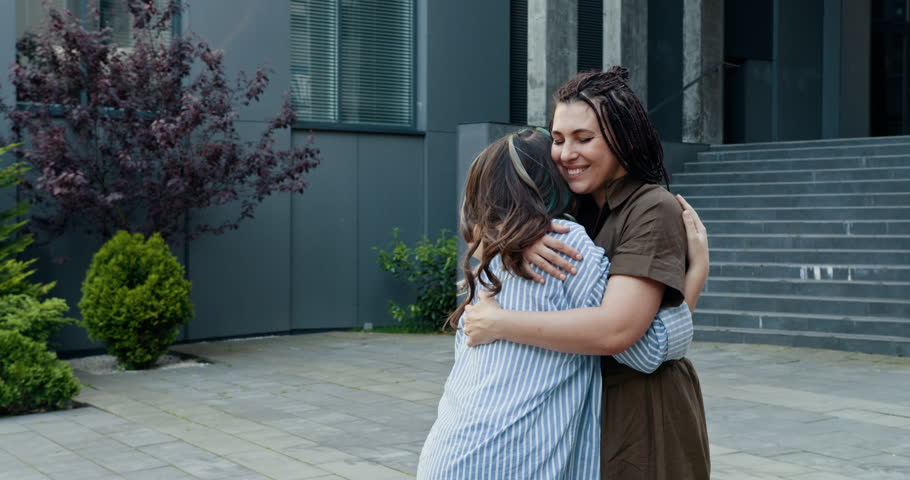 Two women of diverse backgrounds embracing outside modern building, expressing joy and friendship. Middle-aged white woman and young black woman sharing happy moment together.