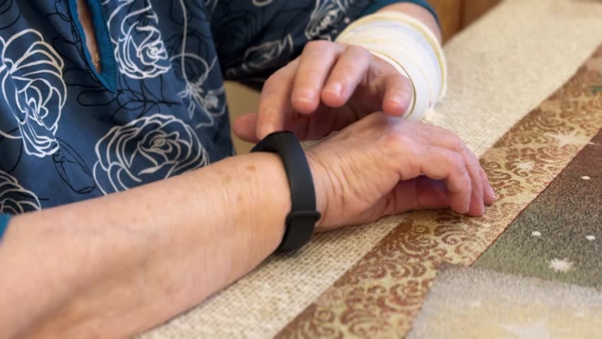 The black smart band with display touchpad and heart rate monitor. The old woman with broken arm with cast for the arm sitting at sofa touches and looks at the wristband of pulse monitor indoors