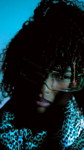 Stylish african american model striking bold poses wearing futuristic colorful glasses against vibrant blue studio backdrop, embodying contemporary cyberpunk aesthetic