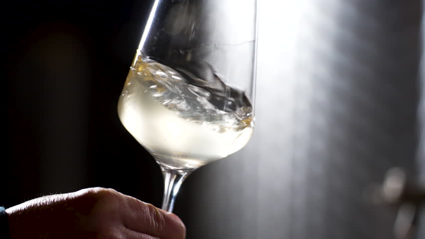 White wine is being swirled in transparent glass by winemaker for flavor tasting. Slow motion close up.