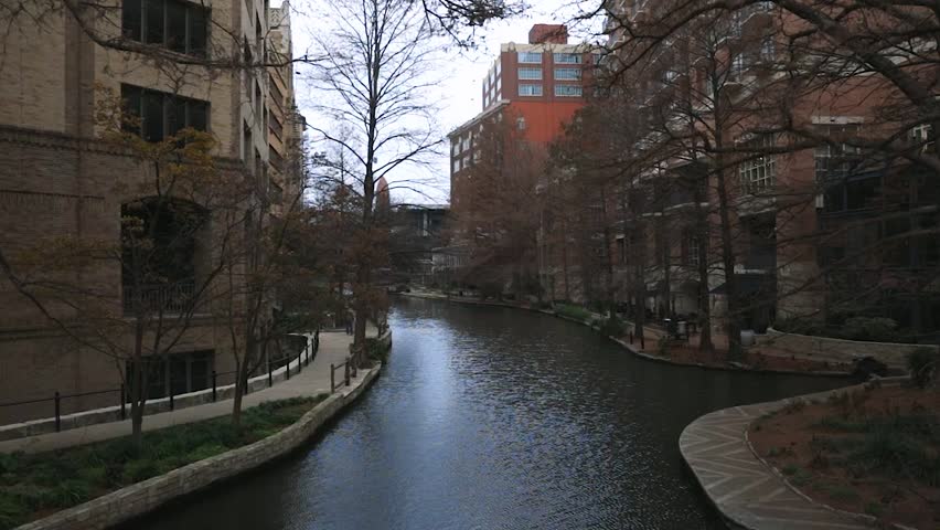 A smooth motion shot glides along an empty river, its calm waters flowing through the heart of the city, flanked by towering buildings and quiet streets