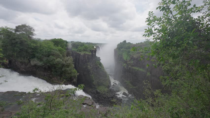 Wide angle of the natural wonder Victoria Falls with a small stream flowing into the mist from the large waterfall that is surrounded by green bush of the Zimbabwian rive banks