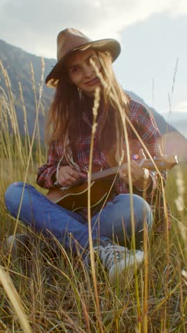 Young woman playing ukulele in a peaceful landscape with majestic mountains and tall grass. Female enjoying lovely music in a charming, serene outdoor setting