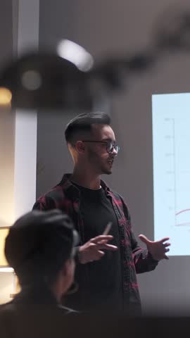 Late At Night Team Of Young Business People Hold a Meeting. A Man Speaks And Uses a Digital Projector For Presentation To a Group Of Colleagues. Growth Graph On Screen