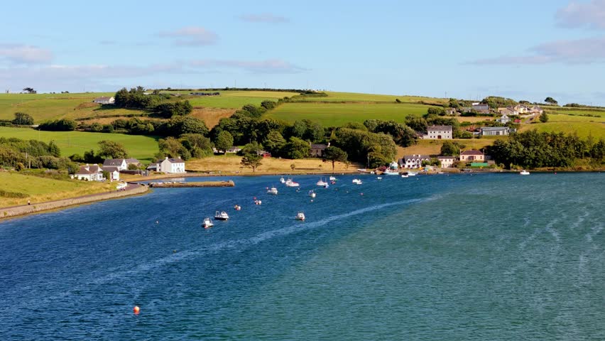 Coastal houses line a calm bay. Numerous small boats are moored in the tranquil waters. Rolling green hills form a picturesque backdrop.