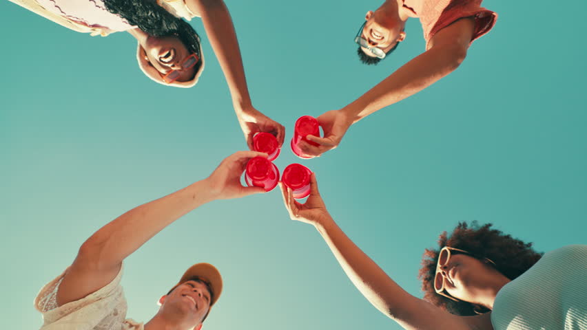 Friends, happy and cheers drinks with celebration for beach party, holiday reunion and summer adventure. Low angle, blue sky and people in circle with alcohol cups for toast, social event and bonding