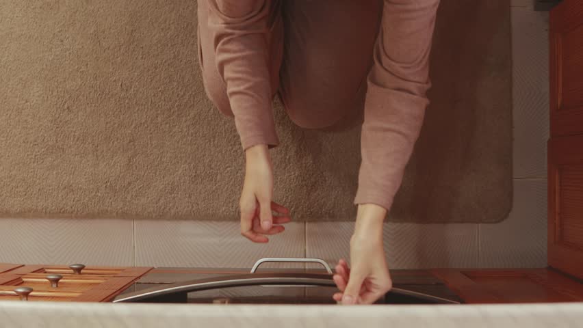 Redheaded woman kneeling in kitchen, wearing casual attire, opening built-in oven door while preparing home meal, revealing dark interior