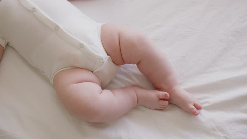 Image in detail of a baby lying on his back on a white surface, in a white bodysuit. The chubby legs and hands of a healthy baby actively waving. The soft, clean and delicate skin of a child