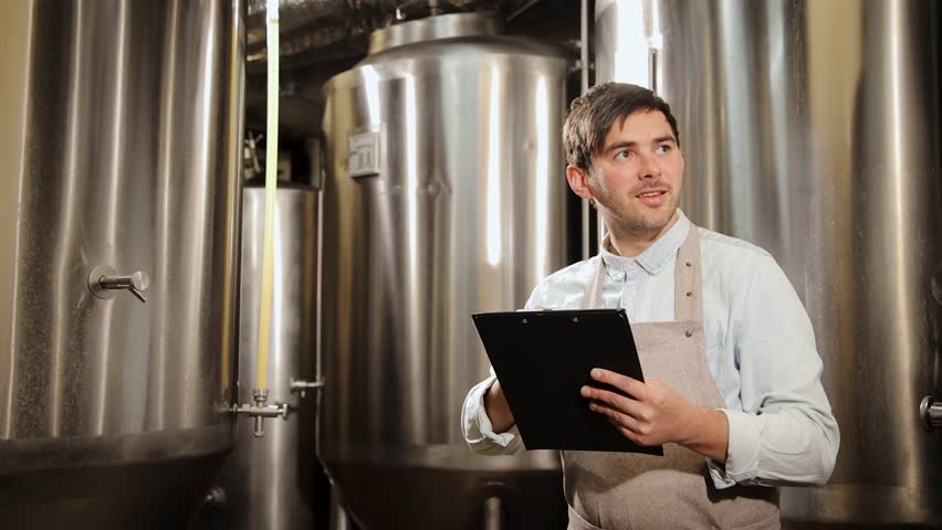 Brewery worker inspecting equipment with clipboard or paper tablet in modern industrial environment