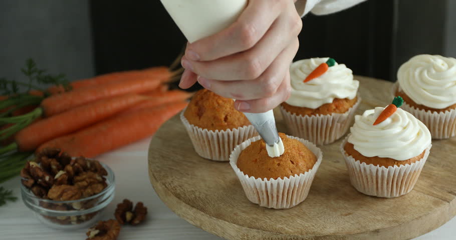 Woman putting cream onto carrot muffin at white wooden table, closeup