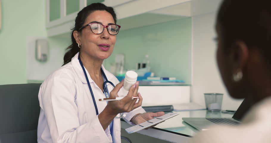Mature doctor talks to young African woman patient, holds bottle with meds, explain how to take pills, gives medication prescription paper at professional consultation at hospital. Treatment, pharmacy