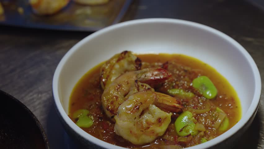 A closeup slow-motion of human hand with metal tongs, adding one shrimp to a white bowl of Spicy prawn rougaille on table