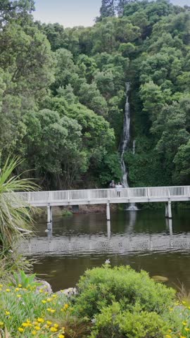 Visitors admire a waterfall cascading down a lush hillside, from a white wooden bridge over a calm river. Nature
