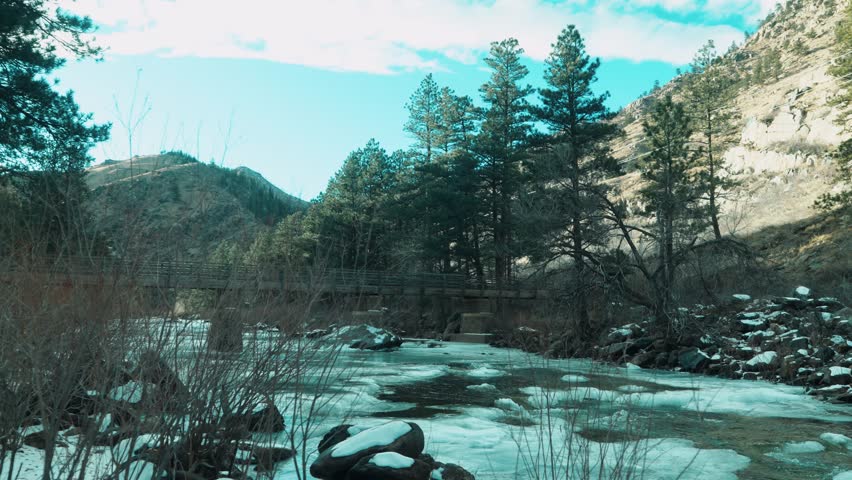 A snow-laden riverbank with flowing winter water, framed by a wooden bridge and rugged Colorado mountain landscape, USA