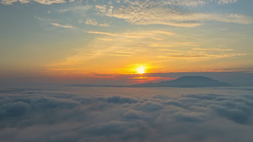 A serene view of the early morning sun rising above a vast sea of clouds, with distant mountain peaks faintly visible on the horizon, creating a peaceful and ethereal landscape. Flying over the cloud