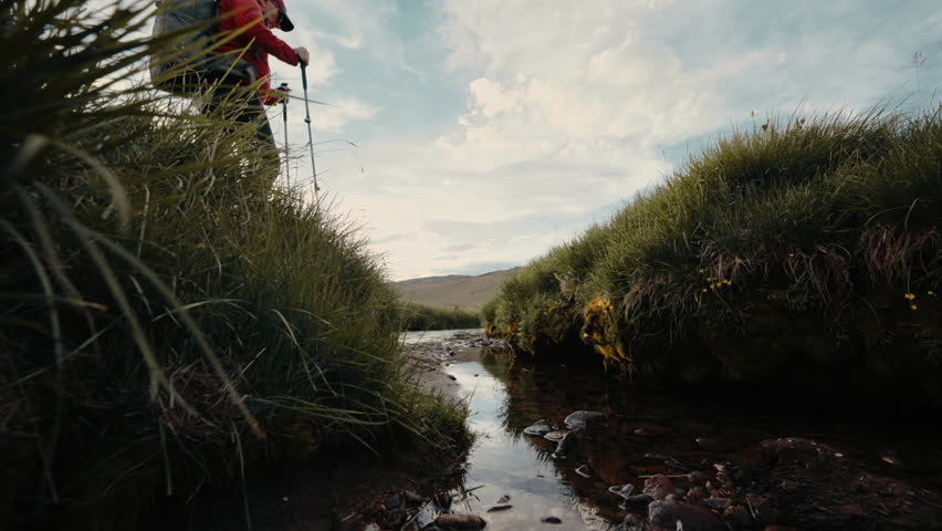 Senior active hiker with backpack and hiking poles is crossing small creek stream