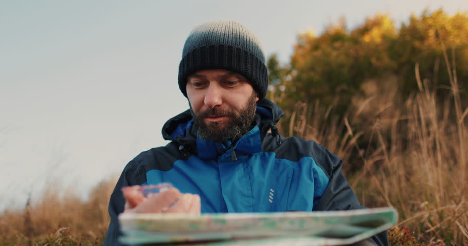 Close-up portrait of a male traveler sitting outdoors, intensely focused on a map and compass. His determined expression and rugged appearance capture the spirit of adventure and exploration as he cha