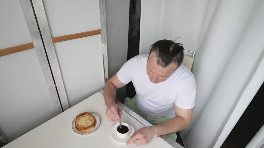A young man drinks aromatic coffee with cottage cheese dessert in an apartment. A cup of black coffee in a man