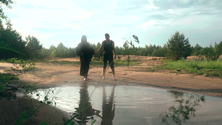 Two individuals walk barefoot beside a tranquil puddle, surrounded by lush greenery and trees. The atmosphere is peaceful, capturing a moment of connection in nature