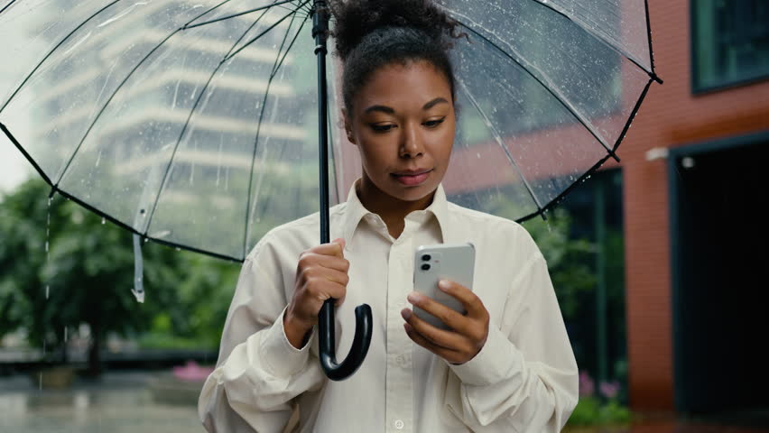African American woman holding using mobile phone smartphone rain female umbrella texting online internet city typing message technology girl businesswoman business chat cellphone connection outdoor