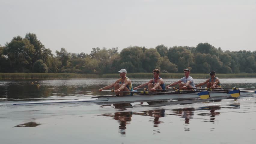 Rowing team training. Side view of 4 young caucasian male rowers, during a rowing practice, athlete sitting in a boat in the river Dnipro, rows through a calm water in autumn. 4k footage. City area in