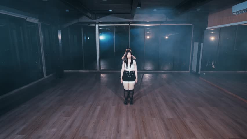 A woman stands in a dimly lit dance studio with wooden floors. The space is empty, with mirrors reflecting her image. Soft blue lighting creates a moody atmosphere.